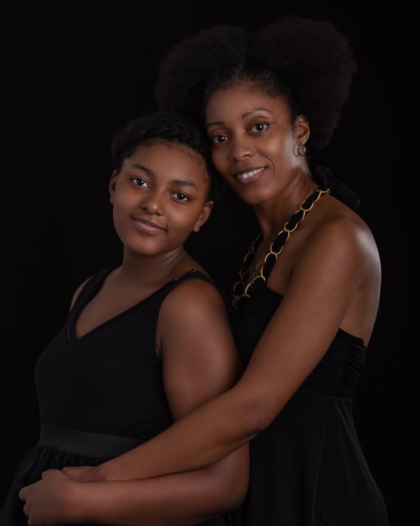 A mother and daughter embracing, both wearing black and posing on a black background